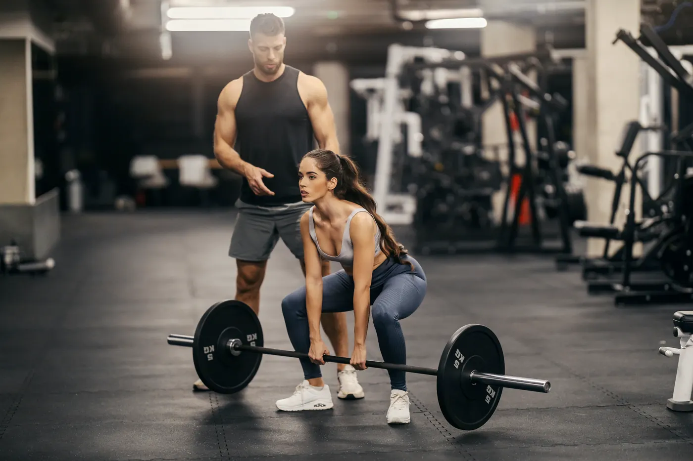 A trainer coaching a woman through a barbell deadlift in a gym